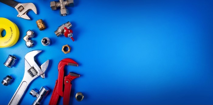 Assorted plumbing tools and fittings on a blue background, including wrenches, pipe fittings, and sealing tape